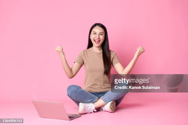young asian woman sitting on floor using laptop and celebrating her success against pink background - fun student stockfoto's en -beelden