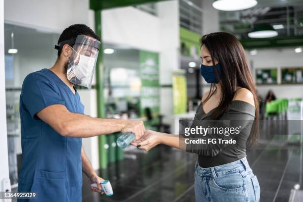 nurse taking temperature and offering hand sanitizer to young woman in line to enter the medical clinic - entering hospital stock pictures, royalty-free photos & images
