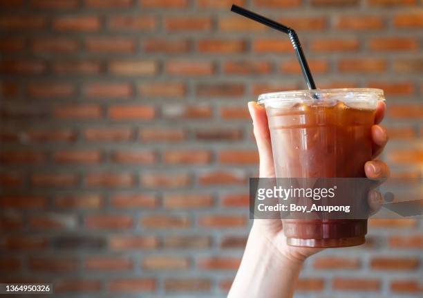 someone hand holding a plastic cup of iced americano (black coffee) against brick wall. - iced coffee foto e immagini stock