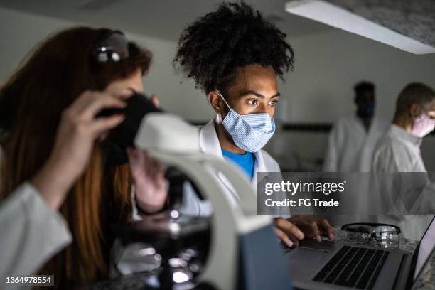 colleagues studying in a lab using laptop and microscopy at university - using face mask - microbiology stock pictures, royalty-free photos & images