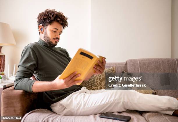 young man reading a book on his sofa at home - hearing aids stock pictures, royalty-free photos & images