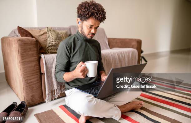 young man having coffee while working on a laptop in his living room - hearing loss at work stock pictures, royalty-free photos & images