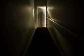A dark stairwell illuminated by a slightly opened door at the top of the stairs.  Shot with a long exposure to create the effect of a sillhouette of a ghost like figure at the top of the stairwell.