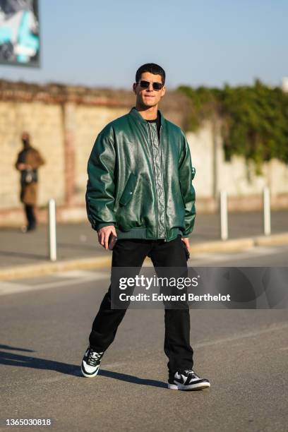 Guest wears sunglasses, a green leather oversized bomber jacket, black pants, black and white Nike sneakers shoes, outside the Prada fashion show...