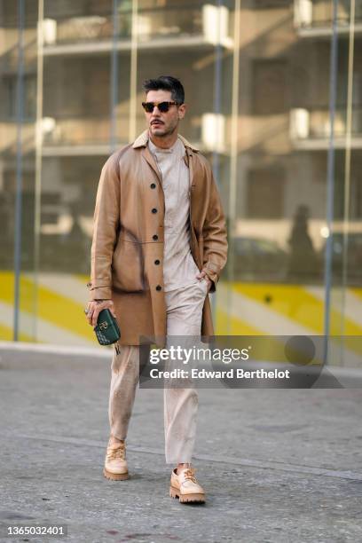 Kadu Dantas wears sunglasses, a black leather coat, a beige floral print sweater, matching pants, a green clutch mini bag, beige and brown shoes,...