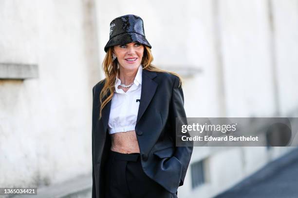 Anna Dello Russo wears a Prada shiny sequined bob hat, a cropped white shirt, a black oversized blazer jacket, matching black suit pants, outside the...