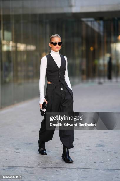 Caroline Daur wears sunglasses, earrings, a white turtleneck top, a sleeveless jacket from a three-piece suit, matching striped black suit pants,...