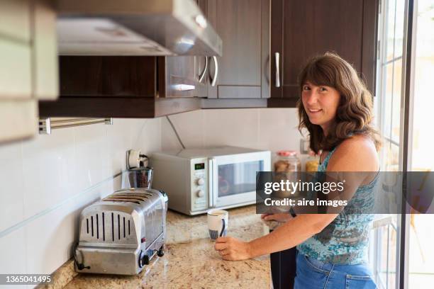 side view of beautiful woman smiling at the camera standing in the kitchen of her home. woman preparing a cup of hot chocolate, sunlight coming through the windows. - microwave dish stock pictures, royalty-free photos & images