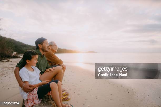 three generation family sitting on beach at sunset - asian grandparents foto e immagini stock