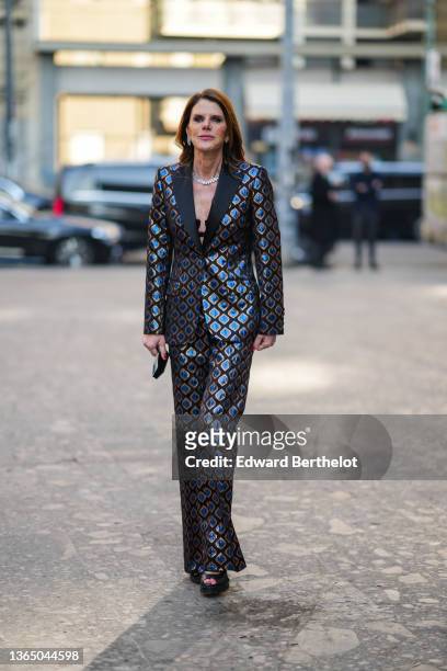 Anna Dello Russo wears a bejeweled necklace, earrings, black bras with floral embroidery, a brown and blue shiny blazer jacket with printed geometric...