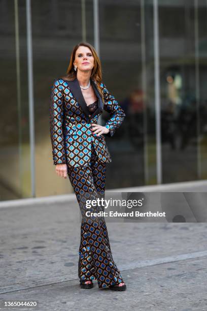 Anna Dello Russo wears a bejeweled necklace, earrings, black bras with floral embroidery, a brown and blue shiny blazer jacket with printed geometric...