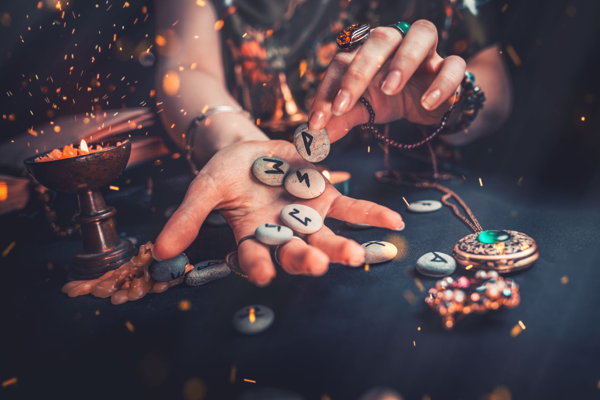 Astrology and esotericism. The witch puts the rune stones in his hand, standing next to the candle. Sparks of fire in the air. Dark background. Astrology and esotericism. The witch puts the rune stones in his hand, standing next to the candle. Sparks of fire in the air. Dark background.