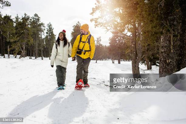biracial young couple holding hands while enjoying a snowshoe walk on a snowy mountain - snowshoeing stock pictures, royalty-free photos & images