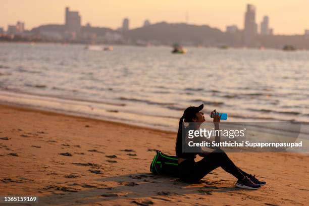 asian woman sitting on the beach and drinking a water for refreshment after exercise during sunset - sport drinking bottle stock-fotos und bilder