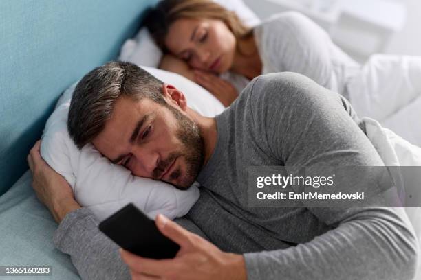 junger mann benutzt handy und checkt nachrichten im bett, während seine frau schläft - ehebruch stock-fotos und bilder