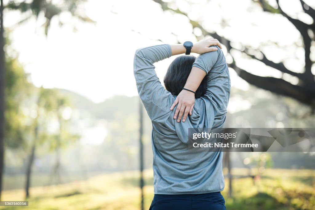 Helps to heal and prevent back pain. Rear View of Asian women jogger doing behind-head triceps stretching on a trail with a fitness tracker at a city park.