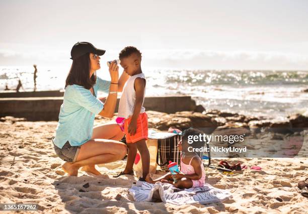 mamá poniéndole loción bronceadora a sus hijos pequeños en la playa - applying fotografías e imágenes de stock