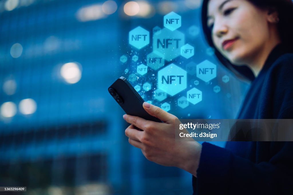 Young Asian businesswoman using smartphone in downtown city street at night, working with blockchain technologies, investing or trading NFT (Non-Fungible Token) on cryptocurrency, digital asset, art work and digital ledger