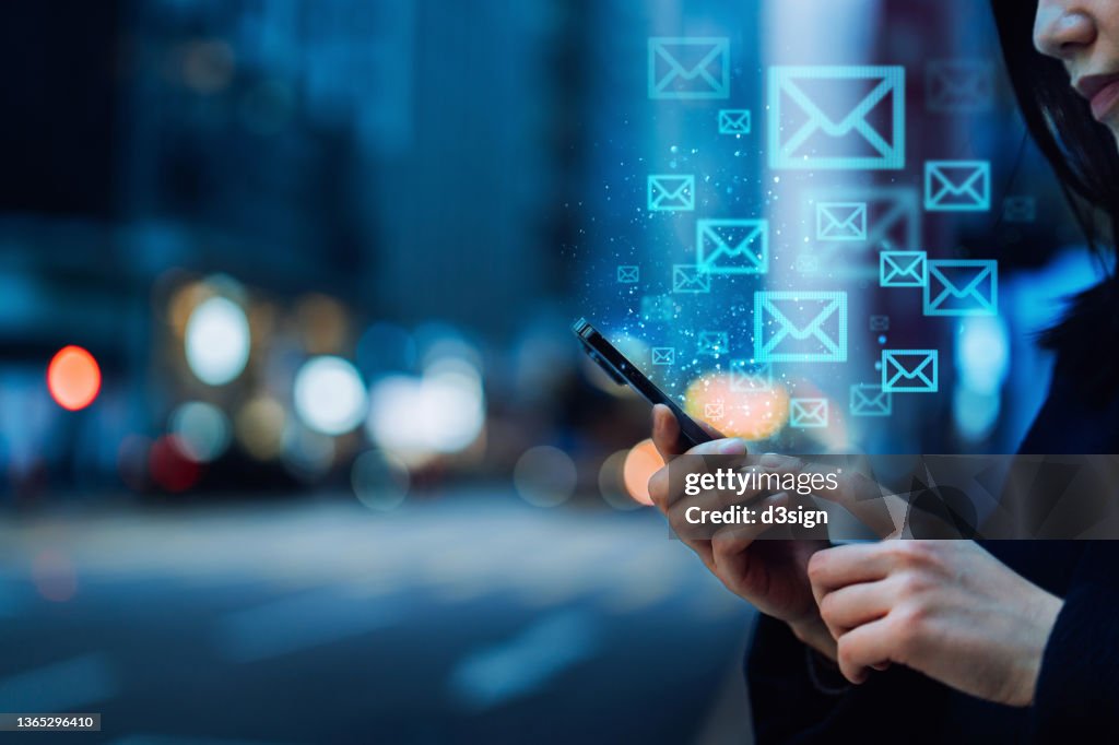 Close up of young Asian woman sending email with smartphone in the city, against illuminated city street lights in downtown district. Information technology concept