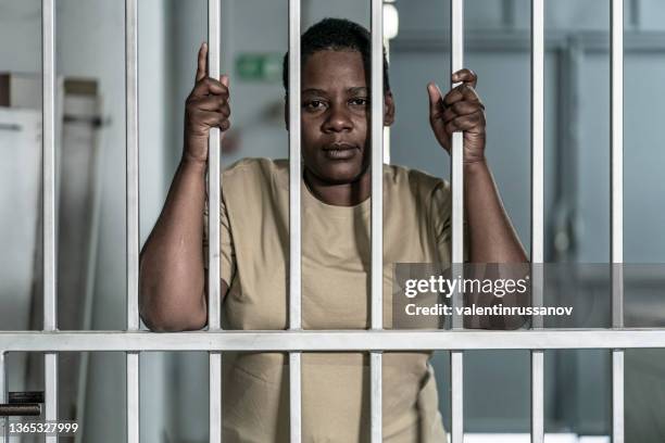 young afro woman looking serious and desperate behind bars which may be prison bars or those of a security gate - woman in jail cell stock pictures, royalty-free photos & images