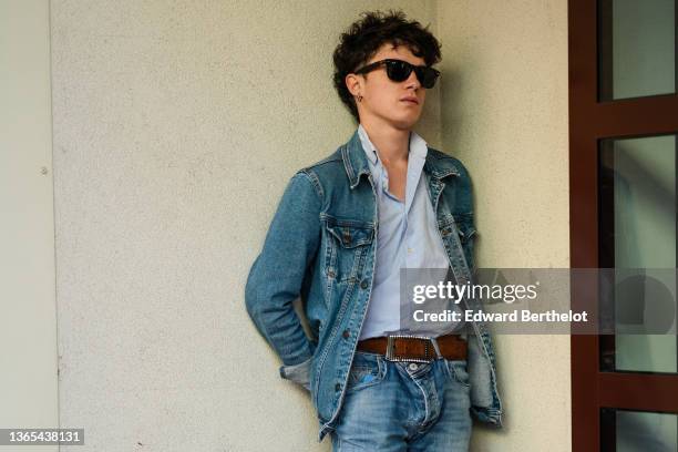 Guest wears sunglasses from Ray Ban, silver earrings, a gray and white small striped shirt, a blue faded denim jacket, a brown shiny leather belt,...