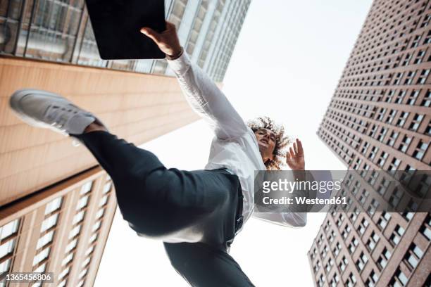 excited businessman holding digital tablet jumping amidst office buildings - blickwinkel der aufnahme stock-fotos und bilder
