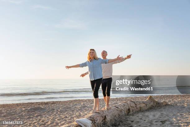 couple with arms outstretched walking on tree log at beach - mature couple winter outdoors stockfoto's en -beelden