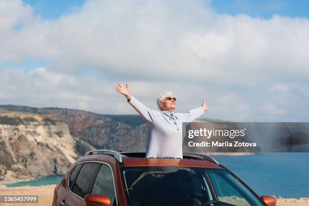 an elderly happy healthy woman on the background of a blue sky smiles, has fun and looks out of the car hatch - longevità foto e immagini stock