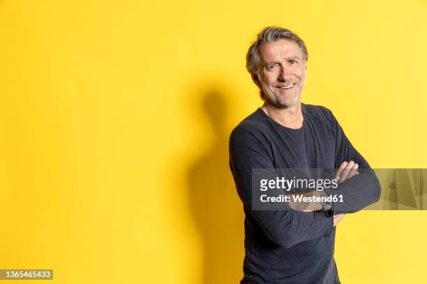 smiling businessman with arms crossed in front of yellow background - dreiviertelansicht stock-fotos und bilder