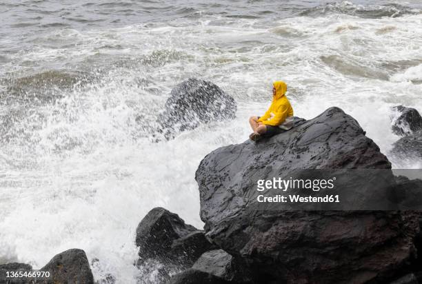 young man wearing raincoat sitting on rock by waves splashing in sea, rocha da relva, san miguel island, azores, portugal - relva stock-fotos und bilder