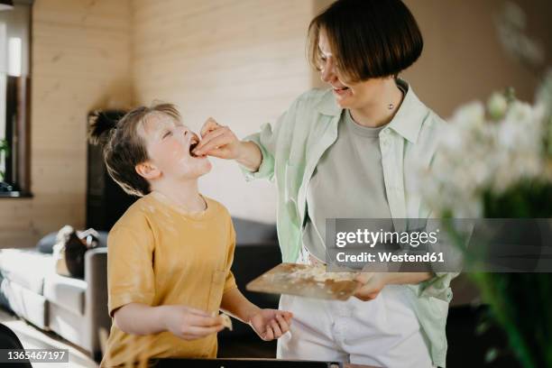 smiling mother feeding grated cheese to son covered with flour on face at home - grated cheese stock pictures, royalty-free photos & images