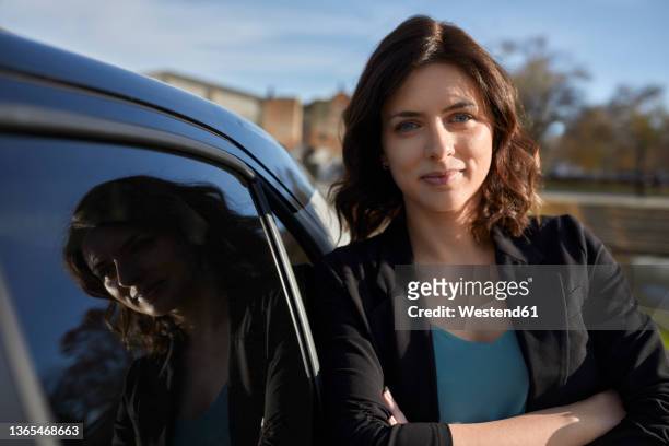 confident businesswoman with arms crossed leaning on electric car - man leaning on car stock-fotos und bilder