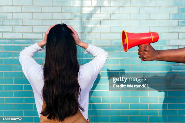 man's hand holding megaphone by woman covering ears in front of turquoise wall - hand to ear photos et images de collection