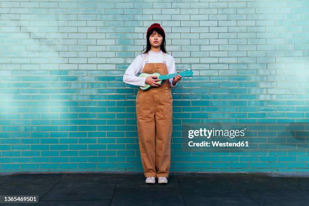 young woman playing ukulele standing in front of turquoise brick wall - ukulele stock-fotos und bilder