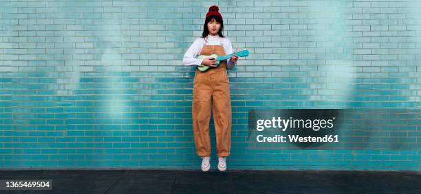 young woman playing ukulele and jumping in front of turquoise brick wall - musician portrait stock pictures, royalty-free photos & images