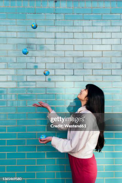 young woman juggling with globes by turquoise brick wall - woman juggling stock pictures, royalty-free photos & images