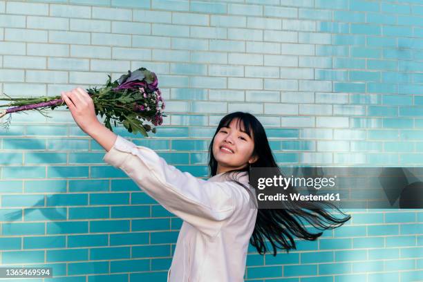 cheerful woman holding bouquet dancing by turquoise brick wall - asian woman long hair stock pictures, royalty-free photos & images