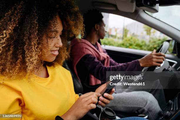 woman using smart phone and friend driving car on road trip - friends inside car stock-fotos und bilder