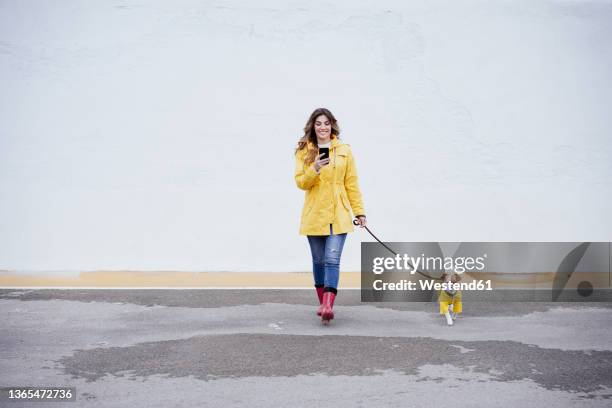 young woman using smart phone and walking with dog on road - matching outfits stock pictures, royalty-free photos & images