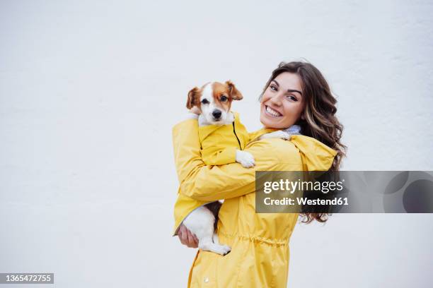 smiling young woman embracing dog by white wall - regnkläder bildbanksfoton och bilder