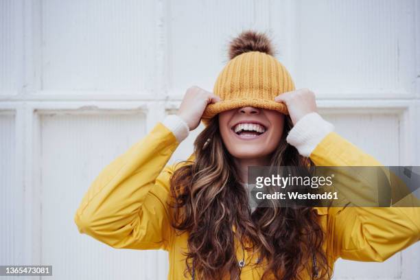 playful woman covering face with knit hat in front of wall - knit hat stock pictures, royalty-free photos & images