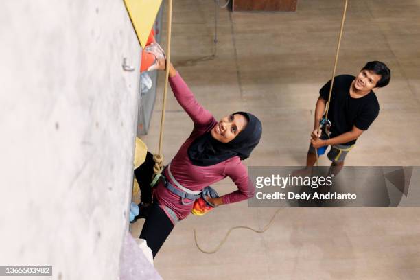 erwachsene muslimische kletterin mit instruktor in indoor-kletterhalle - kletterwand kletterausrüstung stock-fotos und bilder