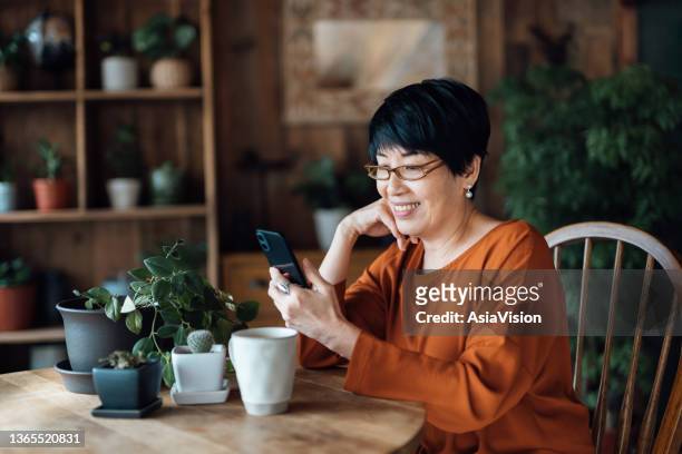smiling senior asian woman managing online banking with mobile app on smartphone, taking care of her money and finances while relaxing at home. retirement lifestyle. elderly and technology - open enrollment begins for third year of affordable care act stockfoto's en -beelden