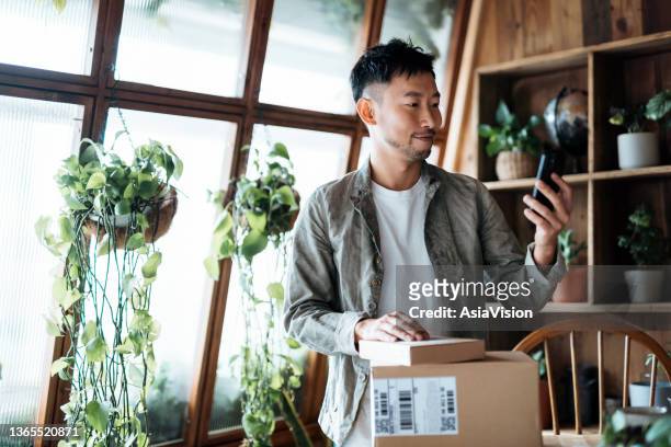 smiling young asian man looking at smartphone while received delivered packages from online purchase at home. online shopping. online banking. shopping and paying safely online - debit cards credit cards accepted stock pictures, royalty-free photos & images