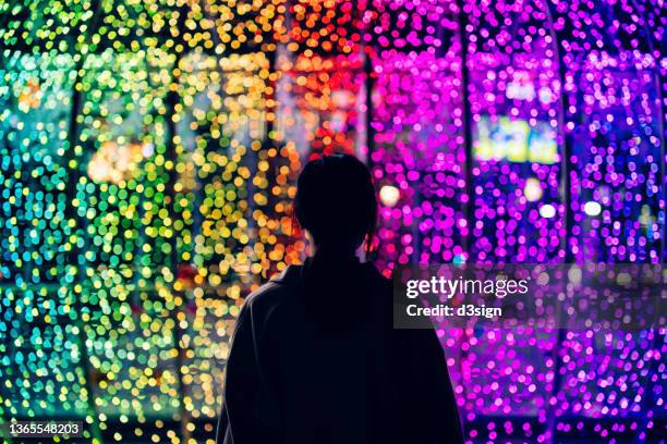 silhouette of young woman standing against illuminated and colourful bokeh lights background in the city at night - christmas scenes stock-fotos und bilder