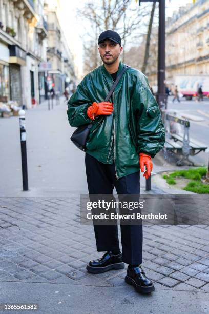 Guest wears black cap, a black t-shirt, a dark green shiny leather oversized bomber coat from Prada, orange shiny leather Prada gloves, a black shiny...