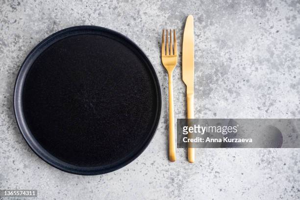 empty black ceramic plate and golden fork and knife on grey concrete background, top view - eetgerei stockfoto's en -beelden