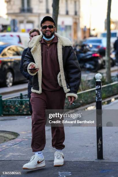 Guest wears a black capblack sunglasses from Balenciaga, a gold large chain necklace, a brown zipper high neck sport sweater, brown sport large...