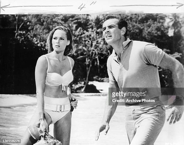 Sean Connery distracted at something he see's while standing near the water at a beach with Ursula Andress in a scene from the film 'James Bond: Dr....