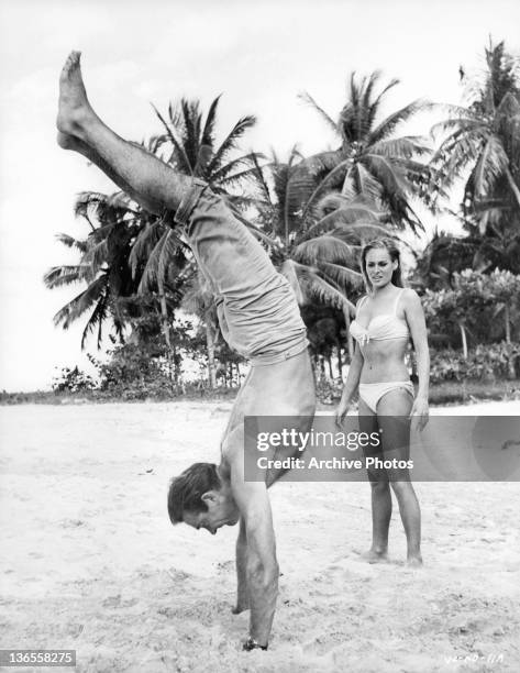 Sean Connery doing a hand stand in the sand while Ursula Andress watches in amazement in a scene from the film 'James Bond: Dr. No', 1962.